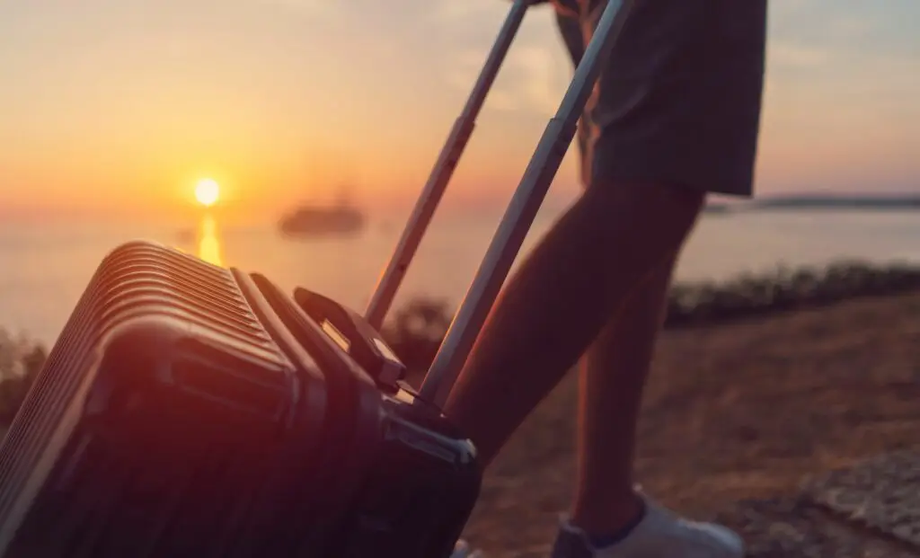 Personne tirant une valise au bord de l’eau au coucher du soleil, symbole de voyage léger et de liberté.